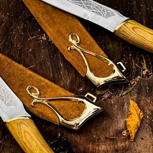May include: Close-up of two knives with decorative blades and wooden handles. The knives are in brown leather sheaths with gold-coloured metal accents. The blades have intricate etched designs.