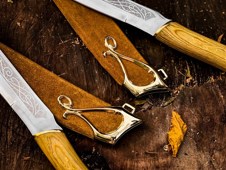May include: Close-up of two knives with decorative blades and wooden handles. The knives are in brown leather sheaths with gold-coloured metal accents. The blades have intricate etched designs.
