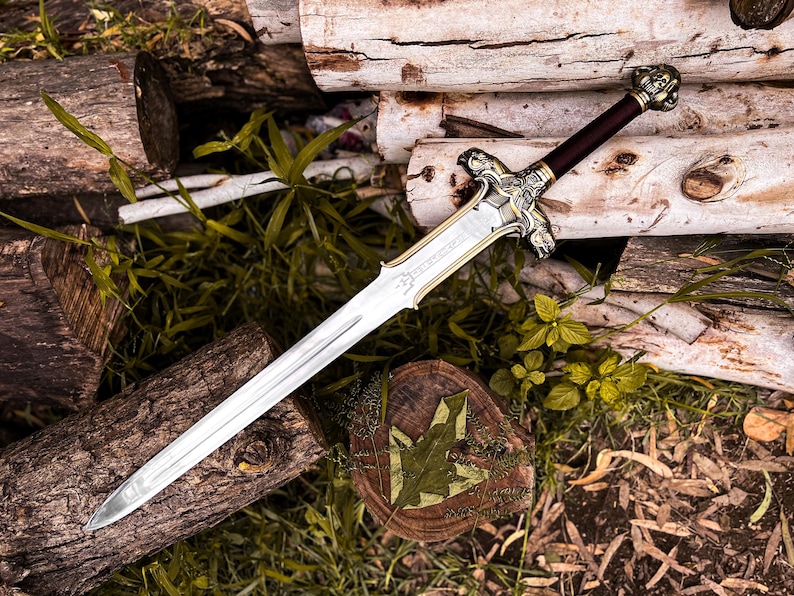 May include: A detailed sword with a silver blade and ornate gold and silver hilt. The handle is dark brown. The sword rests on a bed of wood logs and greenery, creating a rustic and fantasy-themed aesthetic.