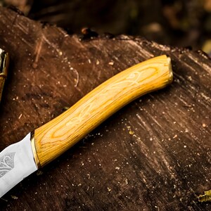 May include: Close-up of a decorative knife. The handle is light wood with a light yellow finish and gold accent. The blade is silver with an etched design. A gold and brown sheath is visible.