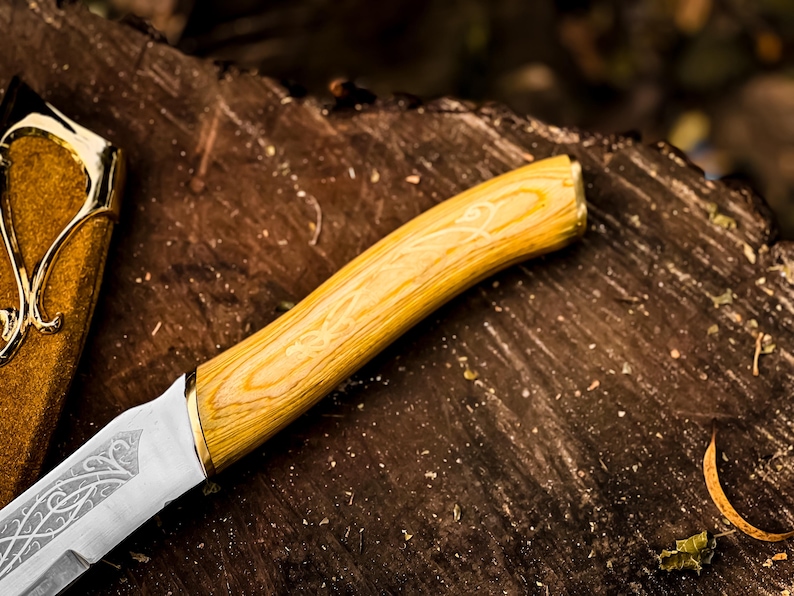 May include: Close-up of a decorative knife. The handle is light wood with a light yellow finish and gold accent. The blade is silver with an etched design. A gold and brown sheath is visible.