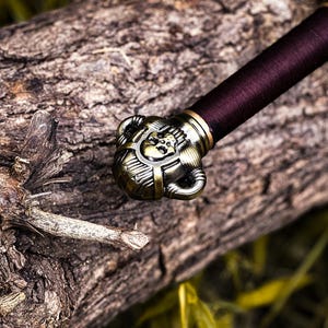 May include: Close-up of a decorative sword hilt and handle. The hilt is ornate with a bronze finish, featuring a skull design. The handle is wrapped in a deep burgundy material. The sword rests on a weathered log.