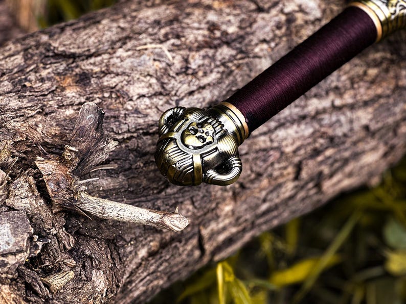 May include: Close-up of a decorative sword hilt and handle. The hilt is ornate with a bronze finish, featuring a skull design. The handle is wrapped in a deep burgundy material. The sword rests on a weathered log.