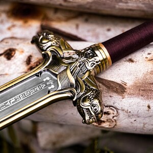 May include: Close-up of a decorative sword with a silver blade and ornate gold-colored hilt. The handle is wrapped in a burgundy material. The hilt features detailed dragon-like designs. The blade has etched inscriptions.