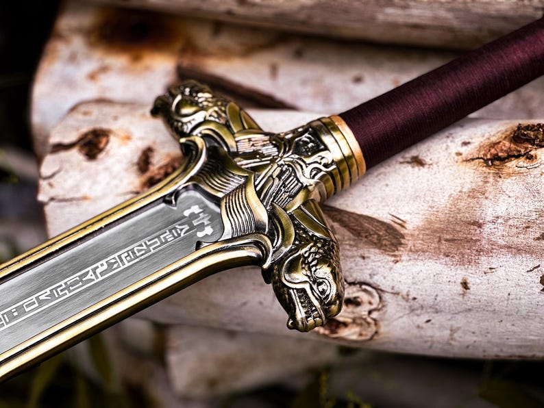 May include: Close-up of a decorative sword with a silver blade and ornate gold-colored hilt. The handle is wrapped in a burgundy material. The hilt features detailed dragon-like designs. The blade has etched inscriptions.