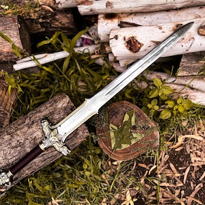 May include: A silver and gold sword with a burgundy handle rests on a bed of logs and greenery. The sword's blade is long and pointed, with intricate detailing on the hilt. The handle is a deep burgundy color, and the sword is placed on a wooden surface.