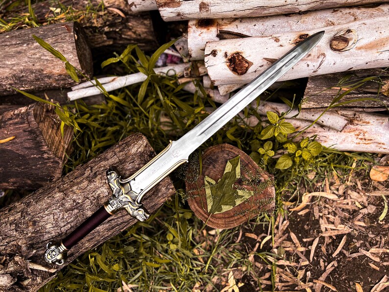 May include: A silver and gold sword with a burgundy handle rests on a bed of logs and greenery. The sword's blade is long and pointed, with intricate detailing on the hilt. The handle is a deep burgundy color, and the sword is placed on a wooden surface.