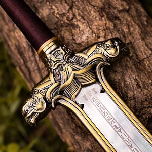 May include: Close-up of a decorative sword with a detailed gold-colored hilt featuring dragon-like figures. The blade is silver with etched designs. The handle is wrapped in a dark brown material. The sword rests on a textured wooden surface.