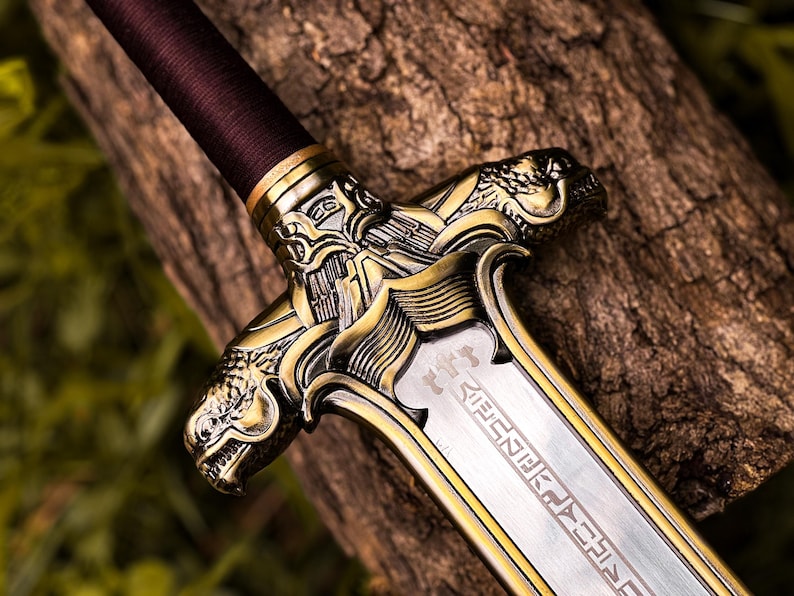 May include: Close-up of a decorative sword with a detailed gold-colored hilt featuring dragon-like figures. The blade is silver with etched designs. The handle is wrapped in a dark brown material. The sword rests on a textured wooden surface.