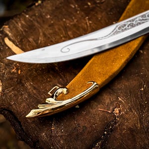 May include: A silver blade with decorative etching rests on a tan leather sheath with gold-coloured accents. The knife and sheath are placed on a rough, brown wooden surface, creating a rustic aesthetic.