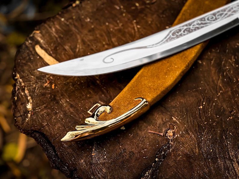 May include: A silver blade with decorative etching rests on a tan leather sheath with gold-coloured accents. The knife and sheath are placed on a rough, brown wooden surface, creating a rustic aesthetic.