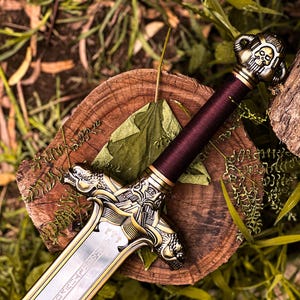 May include: A decorative sword with a silver blade and ornate gold-colored hilt. The handle is wrapped in a deep burgundy material. The sword rests on a wooden surface with a green leaf and foliage.