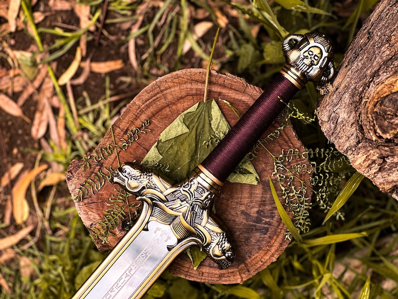 May include: A decorative sword with a silver blade and ornate gold-colored hilt. The handle is wrapped in a deep burgundy material. The sword rests on a wooden surface with a green leaf and foliage.
