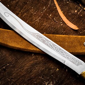 May include: Close-up of a silver blade knife with an ornate etched design. The knife has a wooden handle and a gold-coloured bolster. The knife rests on a brown leather sheath, with a gold-coloured hook.