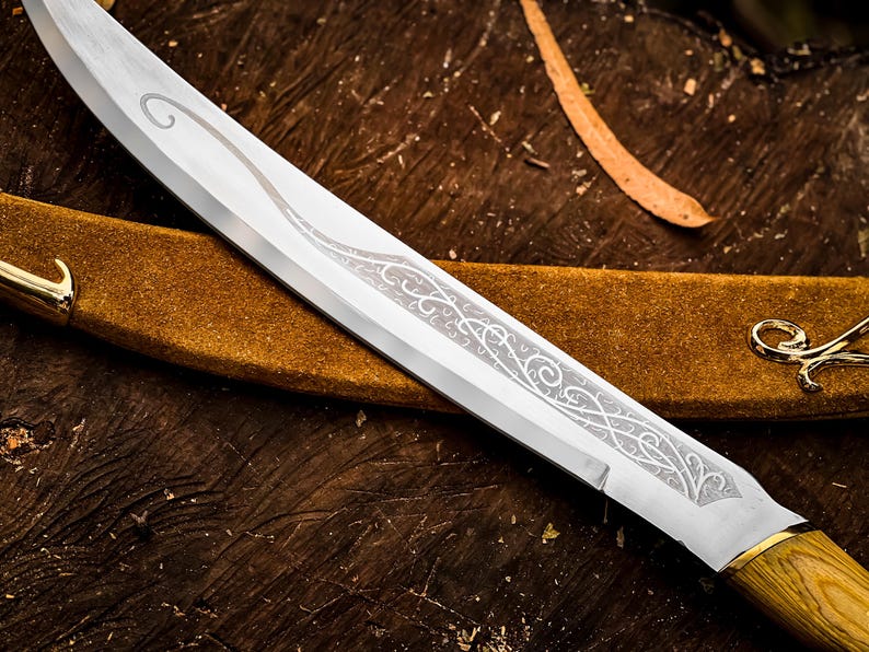 May include: Close-up of a silver blade knife with an ornate etched design. The knife has a wooden handle and a gold-coloured bolster. The knife rests on a brown leather sheath, with a gold-coloured hook.