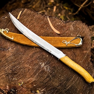 May include: A decorative knife with a light wood handle and a silver blade with etched designs. The knife rests on a wooden surface, with a brown leather sheath and gold-coloured accents.