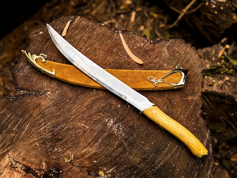 May include: A decorative knife with a light wood handle and a silver blade with etched designs. The knife rests on a wooden surface, with a brown leather sheath and gold-coloured accents.