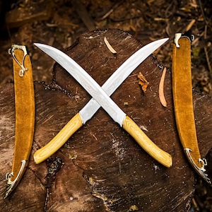 May include: Two crossed curved knives with light wood handles and silver blades rest on a wooden surface. Two brown sheaths with gold accents are on either side. The blades have a unique pattern.