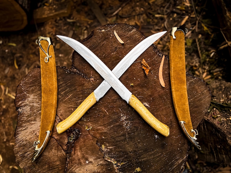 May include: Two crossed curved knives with light wood handles and silver blades rest on a wooden surface. Two brown sheaths with gold accents are on either side. The blades have a unique pattern.