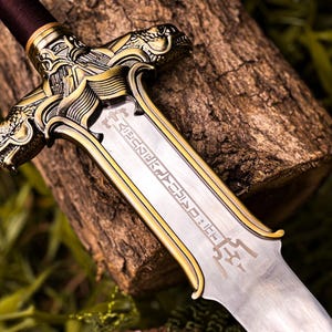 May include: Close-up of a decorative sword with a silver blade and gold-colored accents. The hilt features intricate dragon-like designs. The sword rests on a piece of wood, with green foliage in the background.
