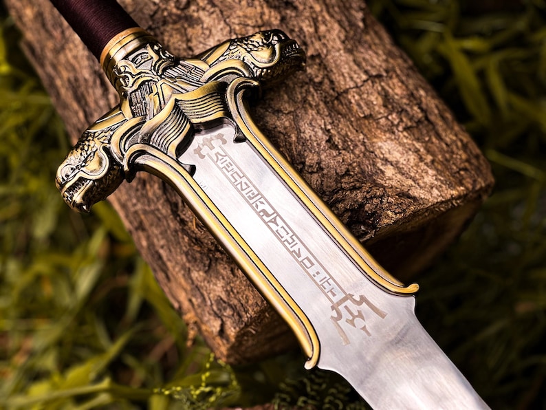 May include: Close-up of a decorative sword with a silver blade and gold-colored accents. The hilt features intricate dragon-like designs. The sword rests on a piece of wood, with green foliage in the background.