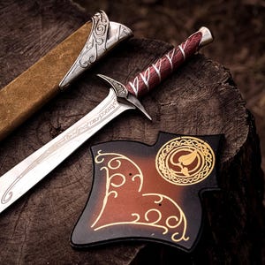 May include: A metal sword with a silver-toned blade featuring etched script, a dark red handle with silver accents, and a tan suede scabbard with ornate silver metal fittings. A dark brown plaque with gold leaf-patterned designs is positioned nearby.