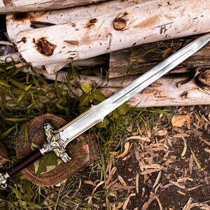 May include: A silver and gold sword with a burgundy handle rests on a wooden surface with greenery. The sword features an ornate hilt and a long, sharp blade. The background includes logs and foliage, creating a rustic aesthetic.