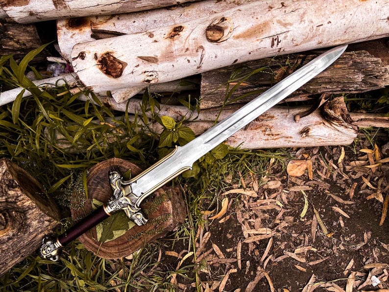 May include: A silver and gold sword with a burgundy handle rests on a wooden surface with greenery. The sword features an ornate hilt and a long, sharp blade. The background includes logs and foliage, creating a rustic aesthetic.