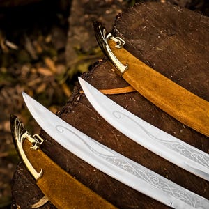May include: Three silver swords with decorative etched blades, gold hilts, and tan suede-like sheaths. The swords are arranged on a dark brown wooden surface. The blades feature intricate patterns.