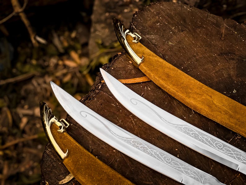 May include: Three silver swords with decorative etched blades, gold hilts, and tan suede-like sheaths. The swords are arranged on a dark brown wooden surface. The blades feature intricate patterns.