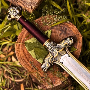 May include: A decorative sword with a burgundy wrapped handle and ornate gold-colored accents. The blade is silver with etched details. The sword rests on a wooden surface surrounded by greenery.