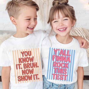 Two young children, a boy and a girl, are smiling and posing together. They are wearing matching white t-shirts with text printed on them.