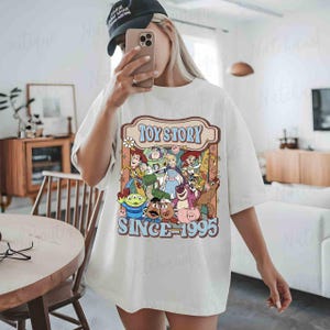 A person is taking a selfie in a room, wearing a white t-shirt with a graphic of characters from the Toy Story franchise.
