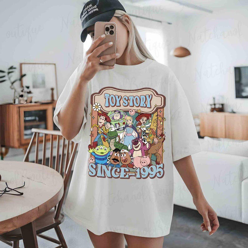 A person is taking a selfie in a room, wearing a white t-shirt with a graphic of characters from the Toy Story franchise.