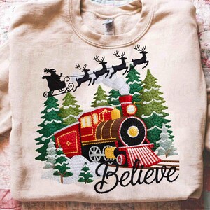 May include: Beige sweatshirt with an embroidered Christmas design. The design features a red and brown train, Santa's sleigh pulled by reindeer, green pine trees, and the word "Believe" in cursive.