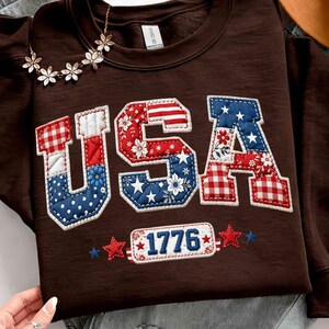 May include: A brown sweatshirt featuring the word "USA" in a patchwork design. The letters are decorated with red, white, and blue patterns, including stars, stripes, and floral prints. The number "1776" is below the letters, with stars on either side.
