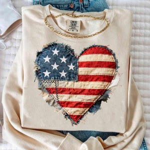 Op de afbeelding: Crèmekleurige sweatshirt met een hartvormig Amerikaans vlagontwerp. De vlag is gemaakt van denim en rood-wit gestreepte stof, met witte sterren. Een gouden ketting ligt op de sweatshirt. Het label luidt "Comfort Colors".