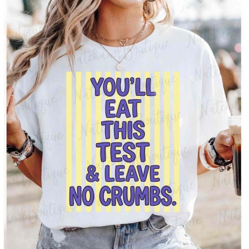 A woman wearing a white t-shirt with a yellow and purple design that says "YOU'LL EAT THIS TEST & LEAVE NO CRUMBS".