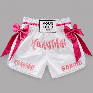May include: White satin Muay Thai shorts featuring a high elastic waistband with a rectangular patch reading YOUR LOGO HERE. The shorts are accented with large hot pink satin bows on each side and pink embroidered text reading MUAY THAI and BOXING.