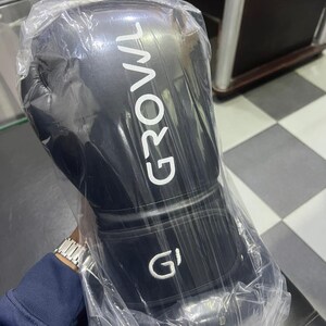 May include: A black boxing glove encased in clear plastic packaging. The glove features the word GROWL printed in white capital letters vertically along the side and a white G logo on the wrist strap. The material appears smooth and matte.