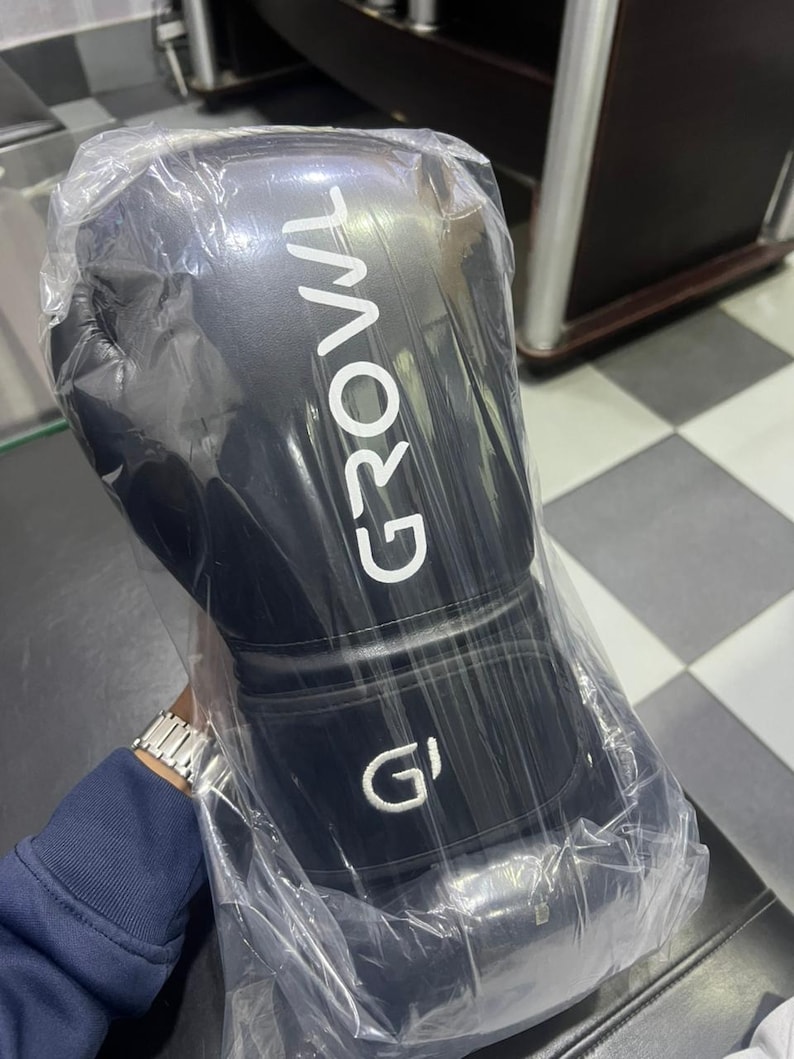 May include: A black boxing glove encased in clear plastic packaging. The glove features the word GROWL printed in white capital letters vertically along the side and a white G logo on the wrist strap. The material appears smooth and matte.