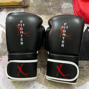 May include: A pair of black boxing gloves featuring white text that reads FIT FIGHTER with a red X graphic. The gloves have a white trim along the wrist strap, which also displays a red X logo. The design is consistent on both gloves.