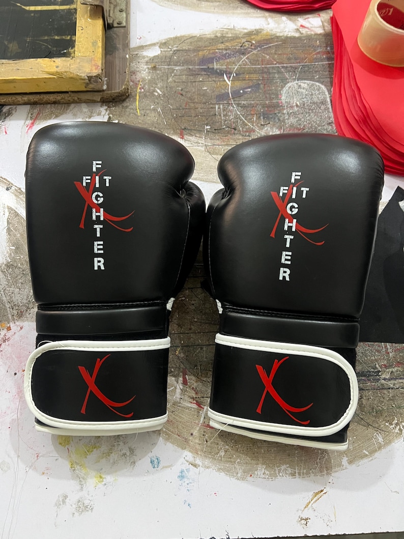 May include: A pair of black boxing gloves featuring white text that reads FIT FIGHTER with a red X graphic. The gloves have a white trim along the wrist strap, which also displays a red X logo. The design is consistent on both gloves.