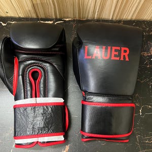 May include: A pair of black boxing gloves with red trim and stitching. The right glove features the word LAUER printed in red on the back of the hand. Both gloves have a wide wrist strap with red piping and white protective paper inserts.