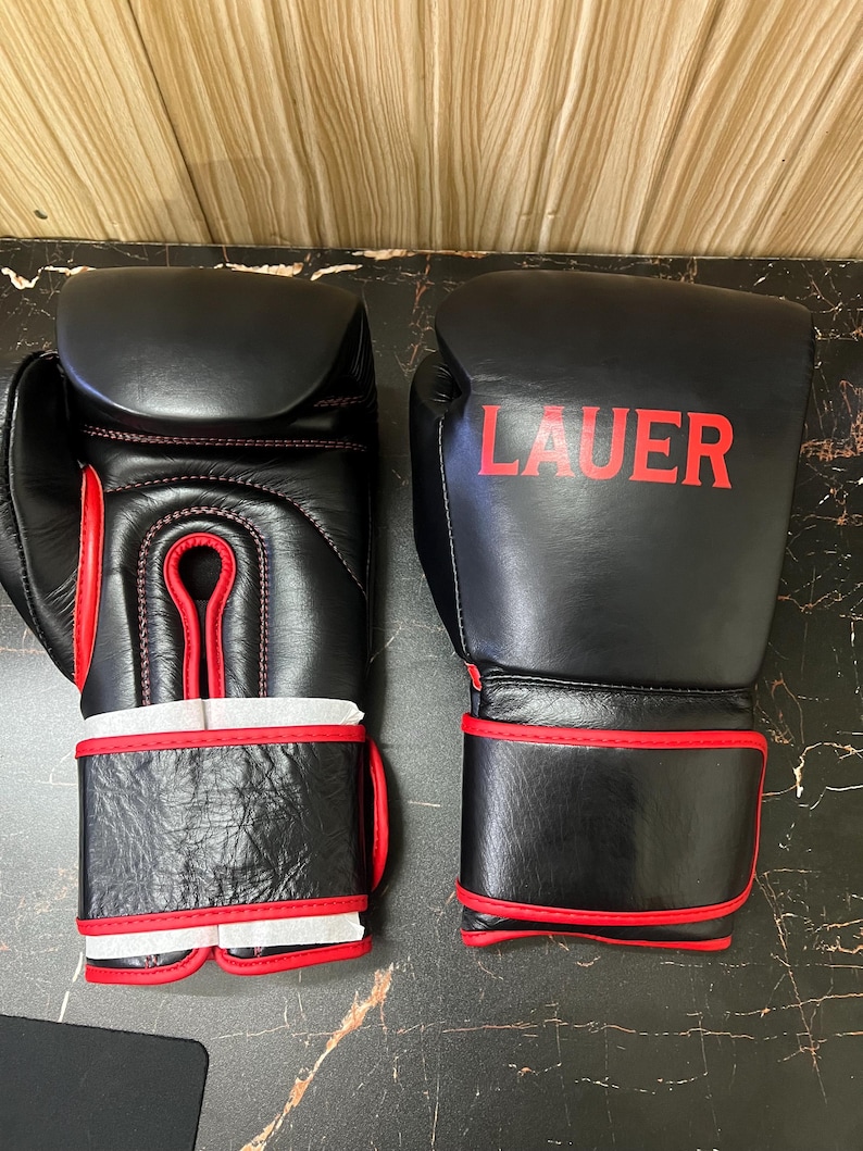 May include: A pair of black boxing gloves with red trim and stitching. The right glove features the word LAUER printed in red on the back of the hand. Both gloves have a wide wrist strap with red piping and white protective paper inserts.
