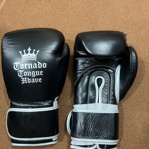 May include: A pair of black boxing gloves with white trim and a white printed logo featuring a crown and the text Tornado Tongue Hdave. The gloves have a smooth finish on the back and a textured leather appearance on the wrist strap area.