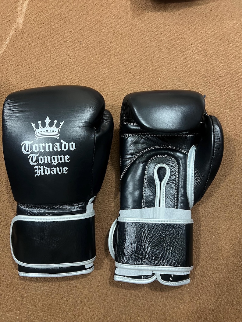 May include: A pair of black boxing gloves with white trim and a white printed logo featuring a crown and the text Tornado Tongue Hdave. The gloves have a smooth finish on the back and a textured leather appearance on the wrist strap area.