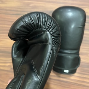 May include: A pair of black boxing gloves featuring a smooth, matte finish and reinforced stitching along the thumb and wrist areas. The gloves have a rounded, padded striking surface and a secure wrist strap closure system for support.