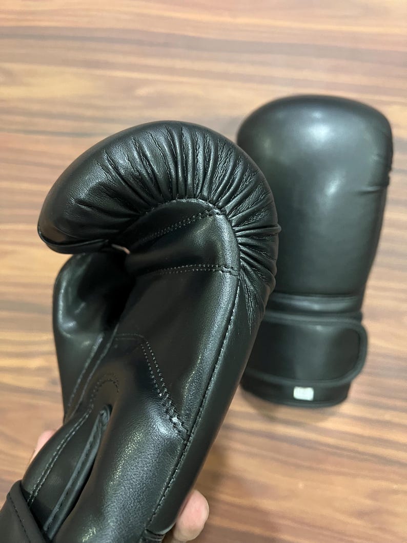 May include: A pair of black boxing gloves featuring a smooth, matte finish and reinforced stitching along the thumb and wrist areas. The gloves have a rounded, padded striking surface and a secure wrist strap closure system for support.