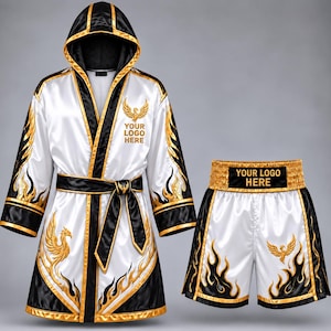 Puede incluir: Una bata de boxeo de satén blanca y negra con capucha y pantalones cortos a juego. Ambas piezas tienen ribetes dorados, patrones de llamas y un emblema de fénix dorado. El texto "YOUR LOGO HERE" aparece en el pecho y en la cintura.